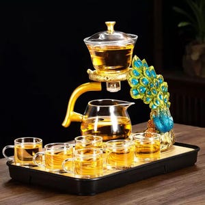 May include: A tea set with a gold-colored stand and a peacock design. The set includes a glass teapot, a brewing vessel, and six small teacups, all arranged on a black tray. The tea is a golden amber color.