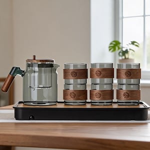 Puede incluir: Un juego de té moderno con una tetera de vidrio con tapa y asa de madera, y cuatro tazas de vidrio con fundas de madera. El juego se exhibe en una bandeja negra con base de bambú. La tetera y las tazas tienen un tinte gris ahumado.