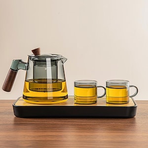 May include: A glass tea set with a teapot, two mugs, and a serving tray. The teapot has a wooden handle and lid. The mugs and teapot are filled with tea. The tray is black with a bamboo insert. The tea set is on a wooden surface.