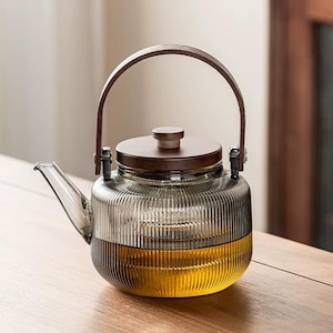 May include: A clear glass teapot with a brown wooden lid and handle. The teapot has a ribbed design and is partially filled with a golden liquid. The handle is made of brown leather and is attached to the glass with metal hardware. A stylish tea accessory.