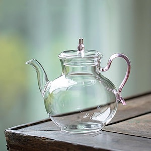 May include: A clear glass teapot with a rounded body, curved spout, and a loop handle. The lid is topped with a small, decorative finial. The teapot is on a wooden surface, with a blurred green background.