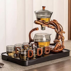 Op de afbeelding: Een theeset met een bruine houten standaard met een aap- en drakenontwerp. De set bevat een glazen theepot, een kan en zes kopjes met houten handvatten. De theeset staat op een zwarte dienblad.