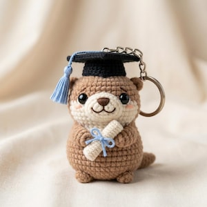 Puede incluir: Un llavero de nutria de ganchillo con un birrete negro y una borla azul. La nutria es de color marrón y crema, sosteniendo un pergamino atado con una cinta azul. El llavero tiene un anillo plateado.