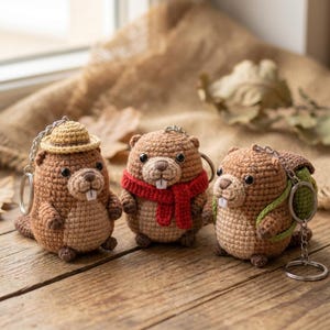 May include: Three handmade crochet beaver keychains. The beavers are brown with black eyes and white teeth. One wears a straw hat, another a red scarf, and the third a green backpack. Each keychain has a silver ring.