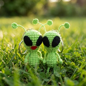 May include: Two green crocheted alien keychains with black eyes and red lips, standing in green grass. Each alien has a silver keychain ring attached. The aliens have antennae with green ball-shaped tips.