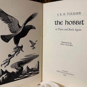 May include: Open book with a black and white illustration of a large eagle carrying figures over a landscape. The text "The Hobbit or There and Back Again" is on the right page, with the author's name and illustrator's name.