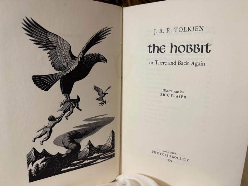 May include: Open book with a black and white illustration of a large eagle carrying figures over a landscape. The text "The Hobbit or There and Back Again" is on the right page, with the author's name and illustrator's name.