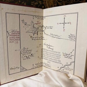 May include: Open book with a detailed map illustration. The map features hand-drawn elements, including a compass, text, and a river. The pages are aged, with a red cover visible. The map is likely from a fantasy novel.