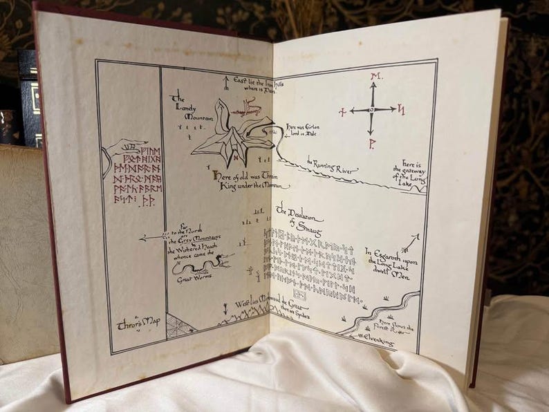May include: Open book with a detailed map illustration. The map features hand-drawn elements, including a compass, text, and a river. The pages are aged, with a red cover visible. The map is likely from a fantasy novel.