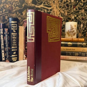 May include: A hardcover book titled "The Hobbit" by J.R.R. Tolkien, with a burgundy cover and gold geometric design. The book is standing upright among other books, with a white cloth in the foreground.