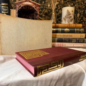 May include: A burgundy book with gold geometric designs on the cover and spine, lying on a white surface. Other books and decorative items are in the background, creating a vintage aesthetic.