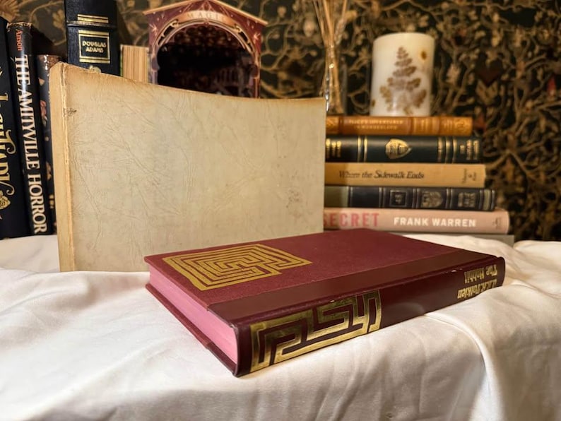 May include: A burgundy book with gold geometric designs on the cover and spine, lying on a white surface. Other books and decorative items are in the background, creating a vintage aesthetic.