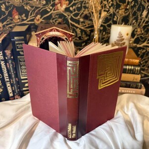 May include: An open, burgundy-colored book with gold embossed labyrinth designs on the cover and spine. The book, titled "The Hobbit" by J.R.R. Tolkien, is displayed with other books and decorative items.