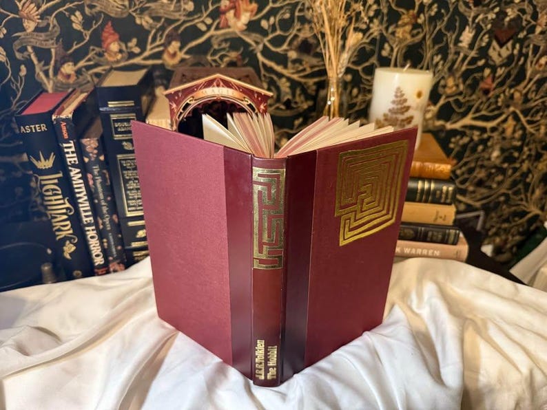 May include: An open, burgundy-colored book with gold embossed labyrinth designs on the cover and spine. The book, titled "The Hobbit" by J.R.R. Tolkien, is displayed with other books and decorative items.