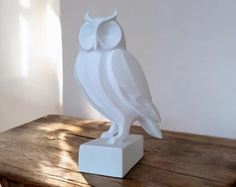 May include: A white owl figurine with a detailed design, standing on a rectangular base. The owl is perched on a wooden table, with a white wall and window in the background. The figurine is a decorative item.