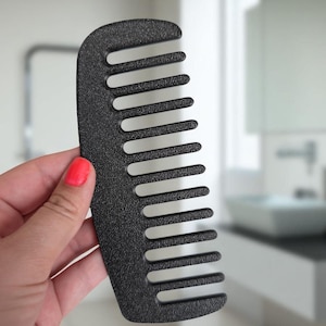 May include: A black wide-tooth comb with a textured surface. The comb has a rounded handle and evenly spaced teeth. The comb is held in front of a blurred bathroom background.