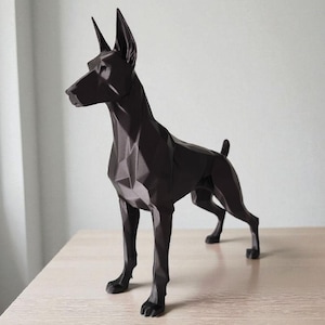 May include: A geometric, black Doberman Pinscher sculpture. The dog is in a standing pose, with its ears erect and tail slightly raised. The sculpture is made of angular planes, giving it a modern, abstract appearance. It is placed on a light-colored wooden surface.