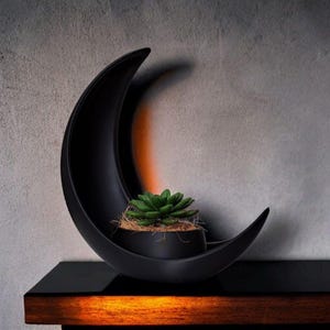 May include: A black, crescent-shaped planter with a small, green succulent inside. The planter sits on a dark surface, with a warm, orange glow visible beneath. The background is a textured, gray wall.
