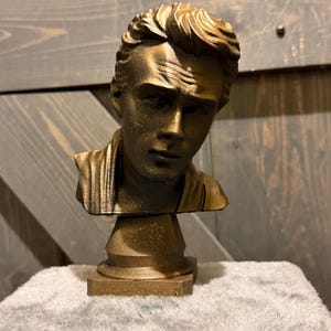 Pode incluir: Uma escultura de busto de cor bronze de um homem com cabelo penteado e uma expressão séria. O busto é montado sobre uma base de pedestal. A escultura é exibida em uma superfície branca e macia contra um fundo de tábuas de madeira.