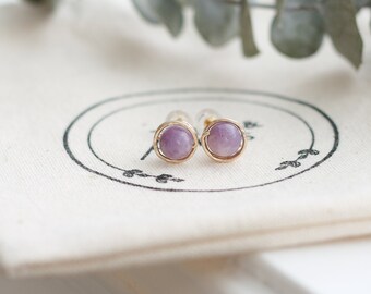 May include: A pair of gold-colored stud earrings with round, light purple gemstones. The earrings are set on a white cloth with a black circular design. Eucalyptus leaves are in the background.