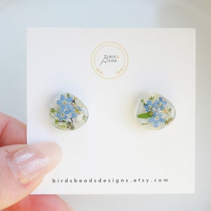 Peut inclure: Une paire de boucles d'oreilles clous avec des fleurs de myosotis pressées dans de la résine transparente. Les boucles d'oreilles sont sur une carte blanche avec le texte "birdsbeadsdesigns.etsy.com".