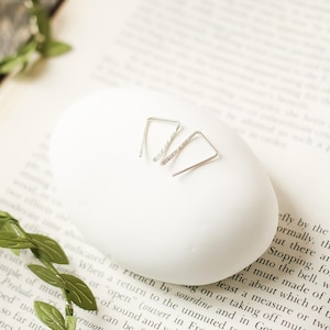 May include: Two silver earrings with a triangular design, one textured and one smooth, displayed on a white oval. The earrings are presented on a book page. The earrings are a modern, minimalist style, suitable for everyday wear.
