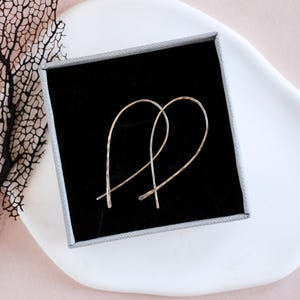May include: A pair of gold-toned metal earrings with a curved, elongated teardrop shape. The earrings feature a smooth, polished finish and are presented inside a square gray gift box with a black velvet interior lining for display.