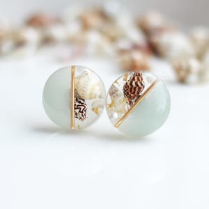 Könnte beinhalten: Ein Paar runde Ohrringe mit hellblauem und goldenem Design. Jeder Ohrring hat einen Abschnitt mit kleinen Muscheln und Goldflocken, die in klarem Harz eingeschlossen sind. Die Ohrringe haben einen vertikalen Goldakzent.