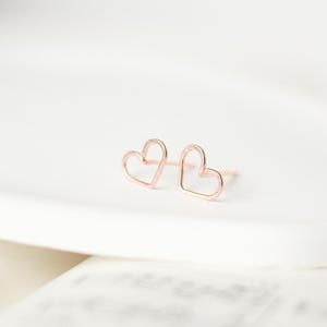 Peut inclure: Une paire de boucles d'oreilles à clous en forme de cœur en or rose. Les boucles d'oreilles ont un design minimaliste. Les cœurs sont soulignés, créant un aspect ouvert. Les boucles d'oreilles sont présentées sur une surface blanche.