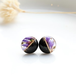 May include: A pair of black stud earrings with a gold trim. Each earring features a clear resin dome with pressed purple flowers.