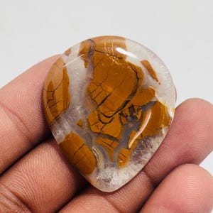 May include: A teardrop-shaped gemstone with a unique pattern of brown and tan veining against a translucent background. The polished stone is held in a hand, showcasing its natural beauty. This stone could be used for jewelry or other crafts.