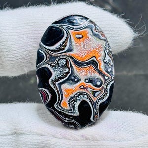 Könnte beinhalten: Ein ovaler Cabochon mit einer glänzenden, marmorierten Oberfläche und wirbelnden Mustern in Schwarz, leuchtendem Orange und Weiß. Der Stein hat ein hochglänzendes Finish mit komplexen, fließenden Schichten, die ein mehrfarbiges, abstraktes Design bilden.