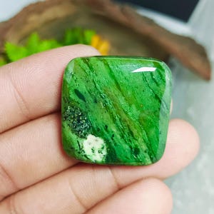 Könnte beinhalten: Ein quadratisch geschliffener, polierter Edelstein in leuchtenden Grüntönen mit einem einzigartigen Muster aus dunkleren grünen Adern und Flecken. Der Edelstein wird in einer Hand gehalten, wodurch seine glatte, reflektierende Oberfläche hervorgehoben wird. Die Ästhetik deutet auf ein natürliches, erdiges Thema hin.