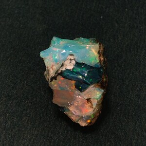 Könnte beinhalten: Ein roher Opal-Edelstein mit einer Mischung aus Farben, darunter Blau, Grün, Orange und Weiß. Der Stein hat eine natürliche, unregelmäßige Form und eine strukturierte Oberfläche. Der Opal ist vor einem dunklen Hintergrund platziert.