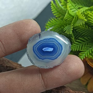 Puede incluir: Una piedra de ágata pulida y redonda con un llamativo patrón de bandas azules y blancas. El centro de la piedra presenta un diseño en forma de ojo azul oscuro, rodeado de anillos concéntricos de azul claro y blanco. Los bordes son ásperos y naturales.