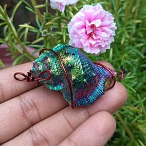 Könnte beinhalten: Handgefertigter Anhänger aus schillernder Muschel mit Drahtwicklung. Die Muschel zeigt ein Farbspektrum, darunter Grün, Blau und Gold. Der Anhänger befindet sich vor einem Hintergrund aus rosa Blüten und grünem Laub.