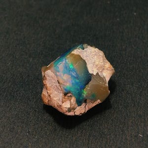Op de afbeelding: Een ruwe, ongeslepen opaal edelsteen met een levendige kleurenpracht. De opaal toont flitsen van blauw, groen en goud in een matrix van bruin en bruin gesteente. De steen is onregelmatig van vorm.