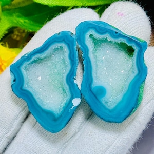 May include: Two polished agate slices featuring vibrant turquoise blue outer bands and a central cavity filled with sparkling white crystalline druzy. The stones exhibit concentric ring patterns in varying shades of blue, highlighting the natural geological formation of the mineral specimens.