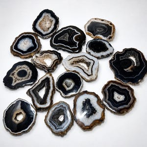 May include: A collection of fifteen polished agate slices featuring concentric bands of black, white, and gray. Each stone displays a unique organic shape with a central geode cavity containing small, sparkling crystals and rough, earthy edges.