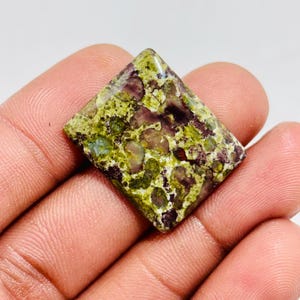 Könnte beinhalten: Ein quadratisch geschliffener Edelstein mit einem gesprenkelten Muster aus Grün, Lila und Braun. Der Stein wird in einer Hand gehalten, was seine einzigartigen Farbvariationen und Textur zeigt. Die Oberfläche des Edelsteins hat ein poliertes Aussehen.