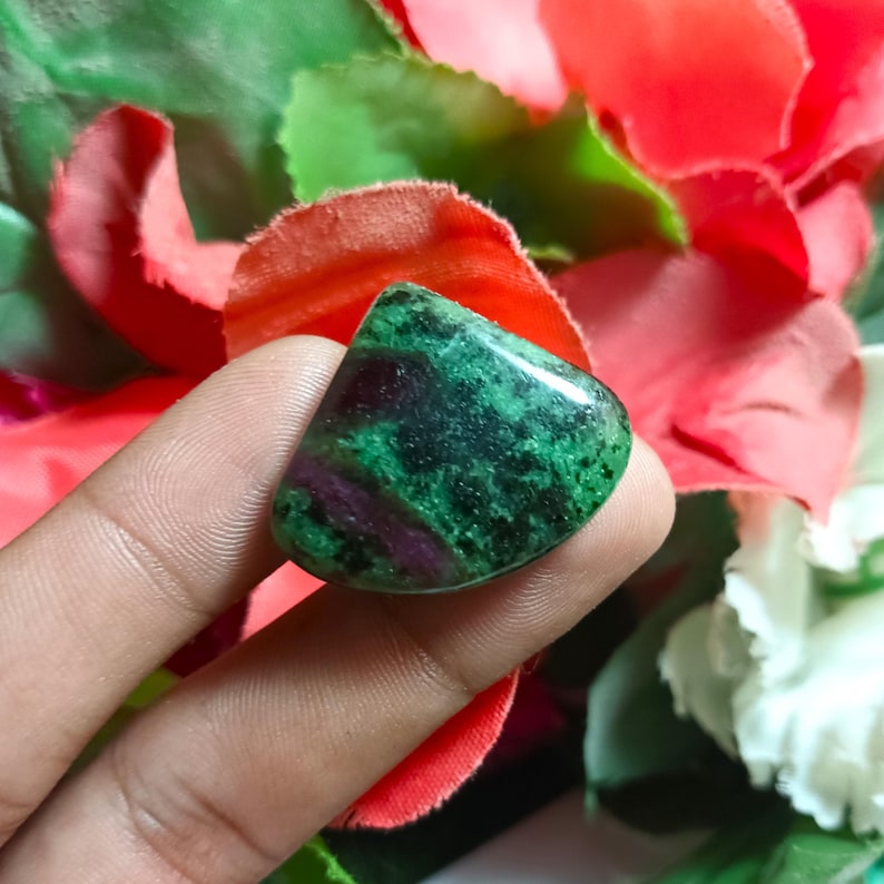 Op de afbeelding: Een gepolijste edelsteen met een mix van groene en dieprode tinten. De steen heeft een gladde, afgeronde vorm en een gespikkeld uiterlijk, waarbij de kleuren in elkaar overlopen. De edelsteen wordt in een hand gehouden, met een achtergrond van rode en witte bloemen.