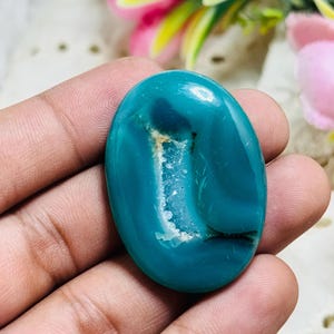 Könnte beinhalten: Ein ovaler, blaugrüner Edelstein-Cabochon mit einer glatten, polierten Oberfläche und einem zentralen, vertikalen, strukturierten weißen kristallinen Einschluss. Der Stein zeigt tiefblaue bis grüne Farbtöne und ein glänzendes Finish.