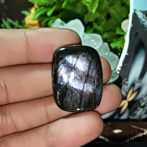 May include: A polished, rectangular labradorite gemstone. The stone has a dark base colour with iridescent flashes of purple and blue. The surface is smooth and reflective, showcasing the natural patterns within the stone. The gemstone is held in a hand.