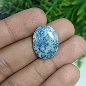Könnte beinhalten: Ovaler Edelstein mit marmoriertem Aussehen. Der Stein weist eine Mischung aus blauen, weißen und schwarzen Sprenkeln auf, die ein einzigartiges Muster bilden. Die polierte Oberfläche reflektiert das Licht und unterstreicht die natürliche Schönheit des Steins. Für Schmuck.