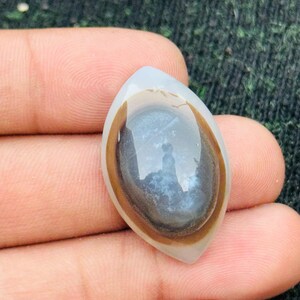 Könnte beinhalten: Ein polierter, tropfenförmiger Edelstein mit einer durchscheinenden, hellfarbigen Außenseite. Die Mitte weist ein dunkles, wirbelndes Muster mit braunen und weißen Bändern auf. Der Stein wird in einer Hand gehalten und zeigt seine natürliche Schönheit.