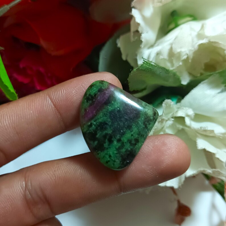 Op de afbeelding: Een gepolijste edelsteen met een mix van groene en donkerrode tinten. De steen heeft een gladde, afgeronde vorm en een gespikkeld patroon. De edelsteen wordt tussen vingers gehouden, met een vage bloemenachtergrond.