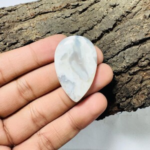 May include: A teardrop-shaped gemstone, polished to a smooth finish, displaying a marbled pattern of white and pale blue. The stone is held in a hand, with a rough, brown wooden surface in the background. Suitable for jewellery.