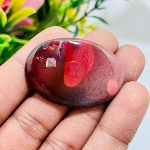 May include: A polished, oval-shaped gemstone with a deep red hue, featuring a lighter red pattern and a small, square, white inclusion. The stone is held in a hand, showcasing its smooth, glossy surface.