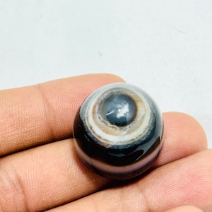 Könnte beinhalten: Ein polierter, runder Edelstein mit einem auffälligen augenartigen Muster. Der Stein weist konzentrische Bänder aus Weiß, Braun und Schwarz auf, die ein einzigartiges, wirbelndes Design erzeugen. Der zentrale Bereich ist dunkelgrau. Der Edelstein wird in einer Hand gehalten.