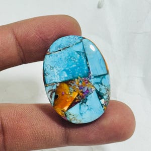 Könnte beinhalten: Oval geformter Edelstein mit einem Mosaikmuster. Der Stein weist eine lebendige Mischung aus Farben auf, darunter Türkisblau, Orange und Lila. Der Edelstein wird zwischen Fingern gehalten und zeigt seine polierte Oberfläche und sein kompliziertes Design.